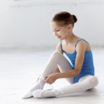 Beautiful little ballerina in blue dress for dancing sitting on the floor and puting on foot pointe shoes on white ballet studio background